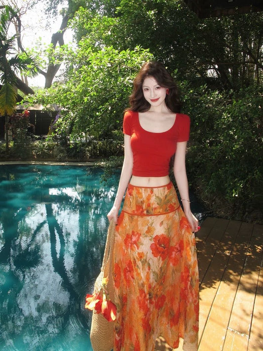 Woman in a red top and floral skirt standing by a pool with greenery in the background